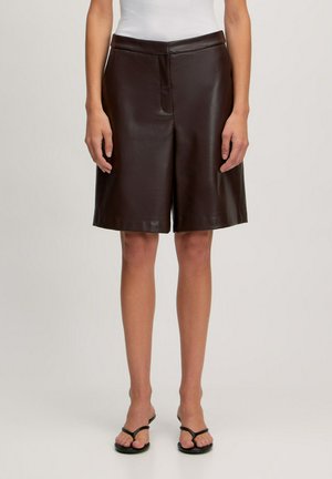Bruine leren shorts met een ontspannen pasvorm, voornaad en tailleband. Gecombineerd met zwarte slippers, weergegeven tegen een neutrale achtergrond.