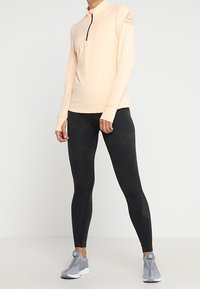 Jasno brzoskwiniowa bluzka z długim rękawem i półzamkiem, zestawiona z czarnymi legginsami o subtelnym wzorze oraz szarymi butami sportowymi.