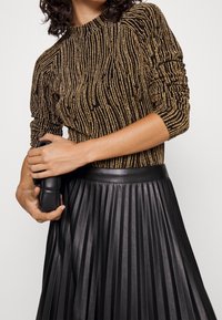 Kvinna i en svart och guldrandig långärmad topp och en svart veckad läderkjol, som håller en liten svart clutchväska.