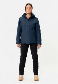 Temno modra vodoodporna jakna z kapuco, z zadrgo spredaj in nastavljivimi manšetami. Nošena čez črne hlače, ki imajo teksturirano tkanino in žepke.