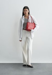 Bolsa vermelha trançada com alças duplas, combinada com uma blusa cinza-claro, calças brancas de perna larga e sapatilhas pretas contra um fundo neutro.