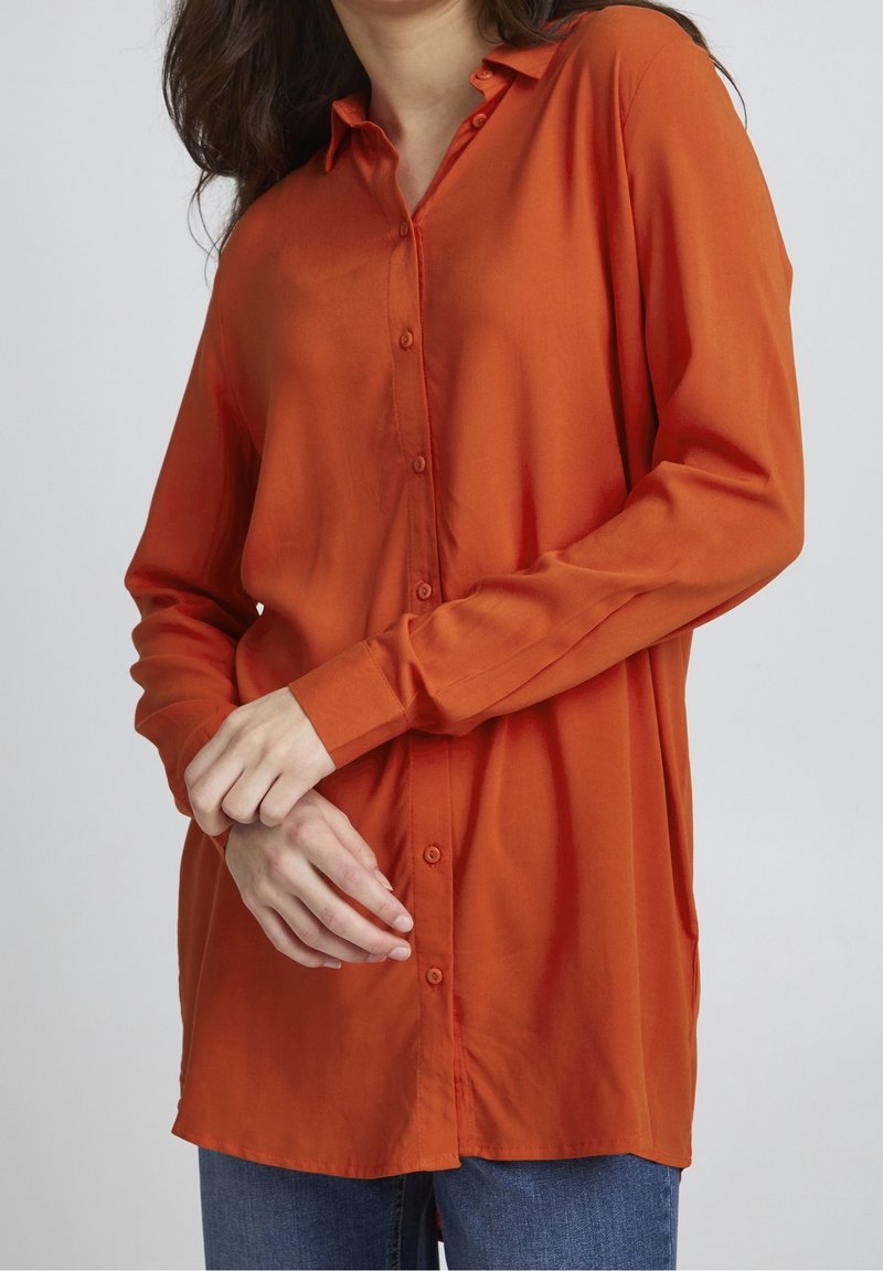 Femme portant une chemise orange à manches longues boutonnée et un jean bleu, les mains doucement croisées à la taille sur un fond uni.