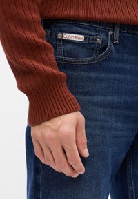 Maglione a coste color ruggine con maniche lunghe abbinato a jeans in denim blu scuro con un logo Calvin Klein ben visibile sulla tasca.
