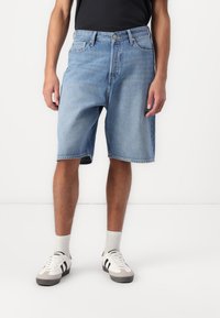Svetlomodré denim šortky s voľným strihom, ktoré majú päť vreciek, zapínanie na gombík a rozstrapkaný lem. Model má na sebe biele tenisky a ponožky.