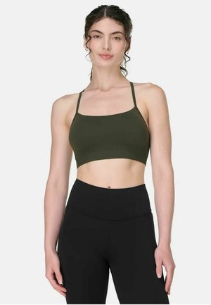 Vrouw met krullend haar, gekleed in een olijfgroene cropped tanktop en een zwarte legging met hoge taille, staand tegen een effen lichte achtergrond.