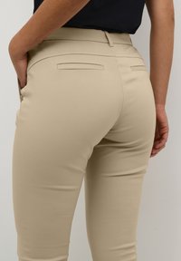Pantaloni beige aderenti con una finitura liscia, dotati di due tasche posteriori e un design a vita media. Dettagli con tasche posizionate a mano.