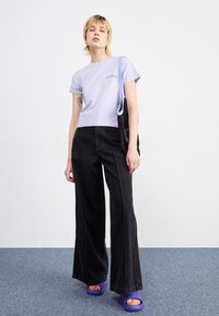 T-shirt in cotone lilla chiaro con maniche corte, abbinato a pantaloni in denim neri a gamba larga e vita alta. Modello con sandali platform viola.