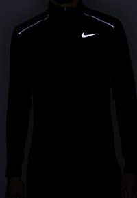 Czarna, długosleeve'owa kurtka sportowa z wysokim kołnierzem, z odblaskowym białym logo Nike oraz paskami biegnącymi wzdłuż ramion. Gładka tekstura.