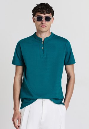 Teal Kurzarmshirt mit Henley-Ausschnitt und drei Holzknöpfen. Glatte Textur, lockere Passform, kombiniert mit weißen Hosen.