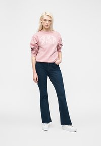 Νεαρή γυναίκα στέκεται φορώντας ροζ φούτερ GAP, σκούρο μπλε τζιν, λευκά αθλητικά παπούτσια, με ένα χέρι στην τσέπη, μπροστά από απλό φόντο.