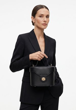 Femme aux cheveux plaqués en arrière portant un blazer noir tient un sac à main structuré en cuir noir avec fermoir et poignée dorés.
