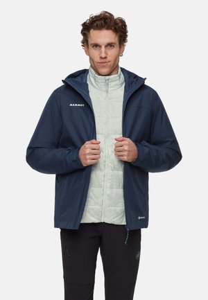 Mann trägt eine marineblaue Mammut Gore-Tex-Jacke über einer hellgrauen, gesteppte Reißverschlussjacke und steht vor einem einfarbigen weißen Hintergrund.