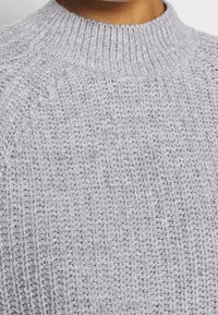 Gros plan d'un pull en maille côtelée gris clair avec un col montant porté par une personne à la peau de ton brun moyen.