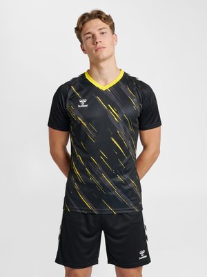 Młody mężczyzna stoi z rękami za plecami, ubrany w czarną odzież sportową z żółtymi paskami i logo hummel na jednolitym białym tle.