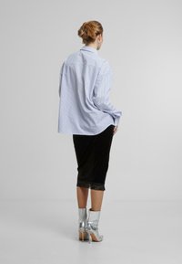 Camicia oversize a righe blu e bianche, gonna nera in velluto con orlo in pizzo e stivaletti metallici argento. Vista posteriore con texture morbida.