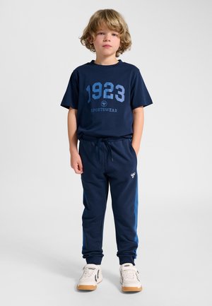 Blond boy standing wearing navy blue sportswear with "1923" on shirt, matching pants, and white sneakers against a plain gray background.