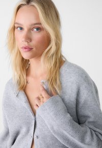 Cardigan grigio a coste con bottoni, caratterizzato da una vestibilità rilassata e una texture morbida. Il modello indossa un anello e mostra un tatuaggio sulla clavicola.