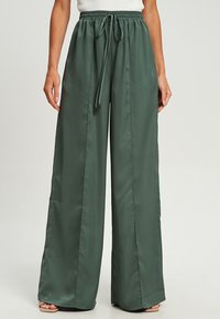 Wijde broek in donkergroene satijnen stof. Elastische tailleband met trekkoord. Glad textuur, vloeiende ontwerp en losse pasvorm.