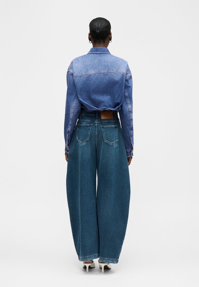 Γυναίκα με μαλλιά σε κότσο, φοράει κοντό τζιν μπουφάν και φαρδιά, ψηλόμεσα τζιν παντελόνια, στέκεται με την πλάτη γυρισμένη σε απλό φόντο.