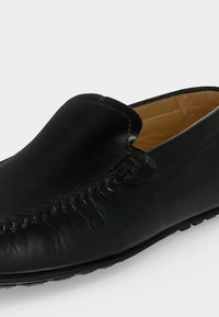 Mocassin noir en cuir sans lacets avec des détails cousus et une doublure intérieure beige sur fond blanc.