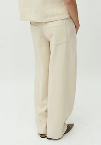 Pantalons beige à jambes larges en tissu texturé, avec deux poches arrière et des coutures latérales visibles, associés à des chaussures noires à bouts pointus.