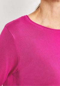 Zbliżenie na osobę w jasnoróżowej, prążkowanej, długiej koszulce z okrągłym dekoltem na neutralnym tle.