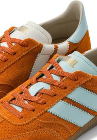 Baskets en daim orange avec des bandes en cuir bleu clair et un talon, languette beige avec texte doré « ANIMAL » et lacets orange.