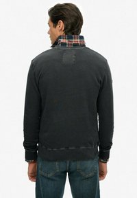 Hombre con sudadera gris oscuro sobre una camisa de cuadros con el cuello levantado, combinado con jeans azules, visto desde atrás contra un fondo liso.