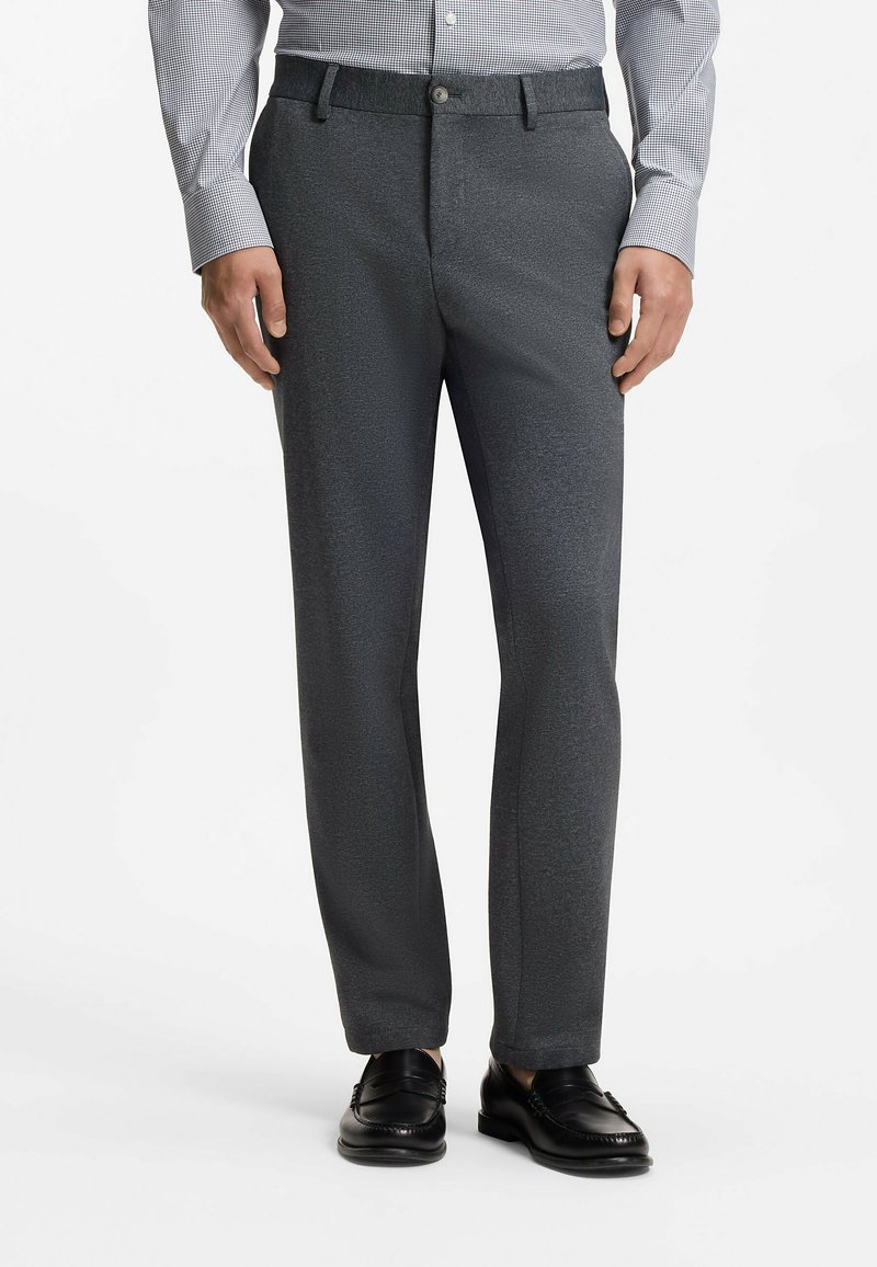 Homme portant un pantalon de costume gris foncé, une chemise à carreaux à manches longues et des mocassins en cuir noirs, debout sur un fond blanc.
