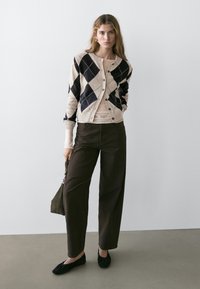 Vrouw staat tegen een witte muur, draagt een beige en zwarte argyle cardigan, beige top, donkere brede broekspijpen, zwarte platte schoenen, en houdt een kleine handtas vast.