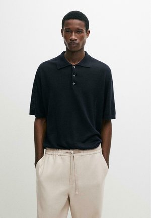 Homme portant un polo noir à manches courtes et un pantalon beige clair avec cordon de serrage, debout devant un fond blanc uni.