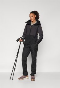 Svart tursjakke med to-toners design, kombinert med svarte bukser. Modellen holder turbuer og har på seg grå og beige sko.