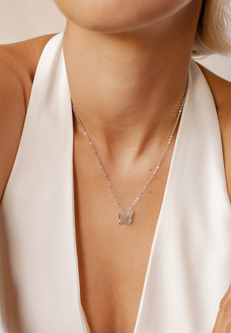 Srebrny naszyjnik z zawieszką w kształcie motyla na delikatnym łańcuszku, noszony z białym ubraniem o głębokim dekolcie, podkreślający skomplikowany design biżuterii.