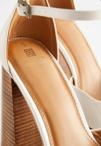Sandales à talons hauts blanches à lanières avec un talon en bois texturé marron. À l'intérieur, une semelle intérieure rembourrée marron clair avec un logo et des détails de taille en relief.