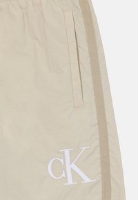 Šviesiai rudos, lengvos šortai su elastine juosta, šoninėmis kišenėmis ir balta siuvinėta "ck" logotipu apatinėje priekinėje dalyje. Tekstūruota medžiaga.
