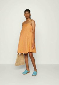 Vestido laranja, texturizado, com alças finas; combinado com uma bolsa tecida e tamancos azuis de madeira. Silhueta simples em A e ajuste descontraído.