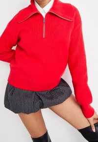 Maglione rosso a maglia con mezza zip sopra una camicia bianca, abbinato a una mini gonna plissettata nera a righe e stivali neri fino al ginocchio.