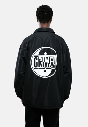 Schwarze Windjacke mit einem großen weißen Yin-Yang und dem Aufdruck "GRIMEY" auf dem Rücken. Sie hat einen kragenförmigen Hals und eine lockere Passform.