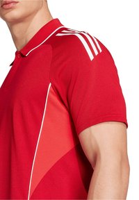 Czerwona koszulka polo wykonana z teksturowanej tkaniny, z białymi akcentami i trzema białymi paskami na ramieniu. Klasyczny design kołnierzyka.