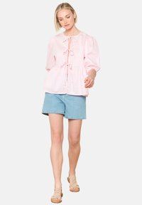 Blusa a scacchi rosa con maniche a palloncino, con un'apertura a chiave davanti e nastri di raso. Abbinata a pantaloncini di jeans chiari e sandali neutri.
