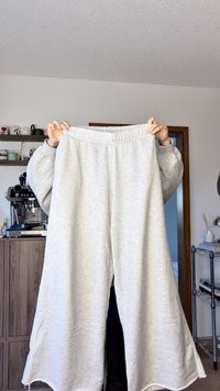 Lichtgrijze sweatpants met brede pijpen en een elastische tailleband, zachte structuur en zonder zichtbare patronen. Vastgehouden door een persoon in een informele setting.