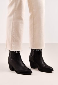 Botas de cuero negras hasta el tobillo con puntas afiladas y costuras decorativas. Tacones gruesos y paneles laterales texturizados. Combinadas con jeans beige de dobladillo deshilachado.