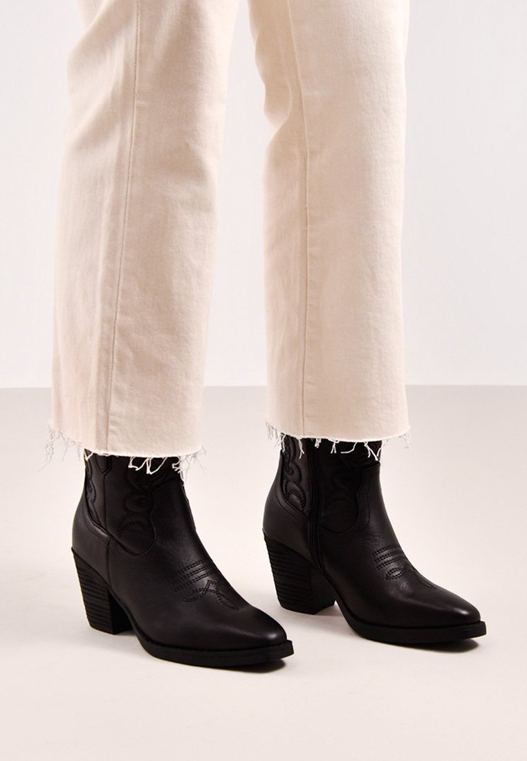 Botas de cuero negras hasta el tobillo con puntas afiladas y costuras decorativas. Tacones gruesos y paneles laterales texturizados. Combinadas con jeans beige de dobladillo deshilachado.