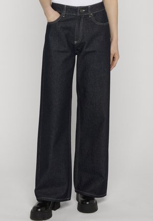 Jeans a gamba larga di colore blu scuro indossati con stivali neri contundenti e una maglia bianca, mani nelle tasche, in piedi contro uno sfondo semplice.