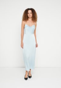 Φόρεμα από σατέν ανοιχτού μπλε χρώματος με εφαρμοστή μπούστα, λαιμόκοψη κασκόλ και λεπτές τιράντες, που τελειώνει λίγο πάνω από τους αστραγάλους. Μαύρα παπούτσια με μυτερή μύτη.