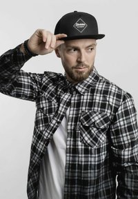 Black and white plaid flannel shirt with two front pockets, layered over a white t-shirt. Black snapback cap with a logo is worn.