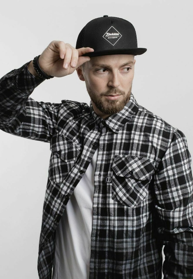 Black and white plaid flannel shirt with two front pockets, layered over a white t-shirt. Black snapback cap with a logo is worn.