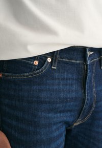Jeans in denim blu scuro presentano una vestibilità slim con cuciture gialle a contrasto, accessori in rame e una chiusura con zip e bottone.