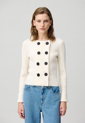 Cardigan bianco a coste con bottoni neri sul davanti, caratterizzato da un collo rotondo e maniche lunghe, abbinato a jeans blu a vita alta.