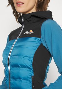 Blå och svart zip-up jacka med vadderad framstycke, huva och logotypdetalj. Materialet har en slät yta med kontrasterande färgpaneler.